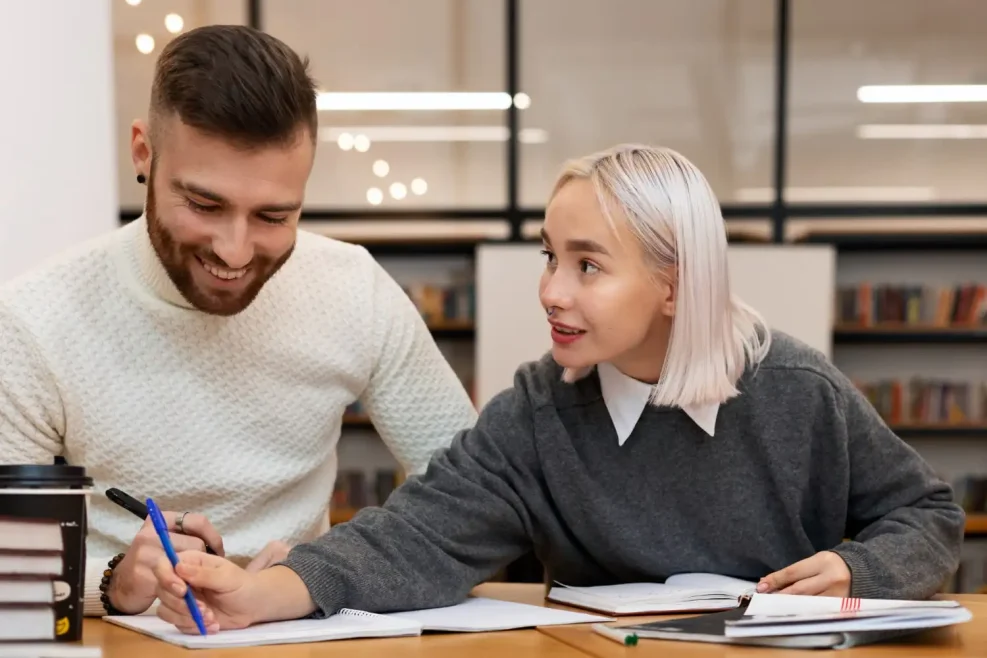 Two students studying English together, discussing idioms and language learning at a library table